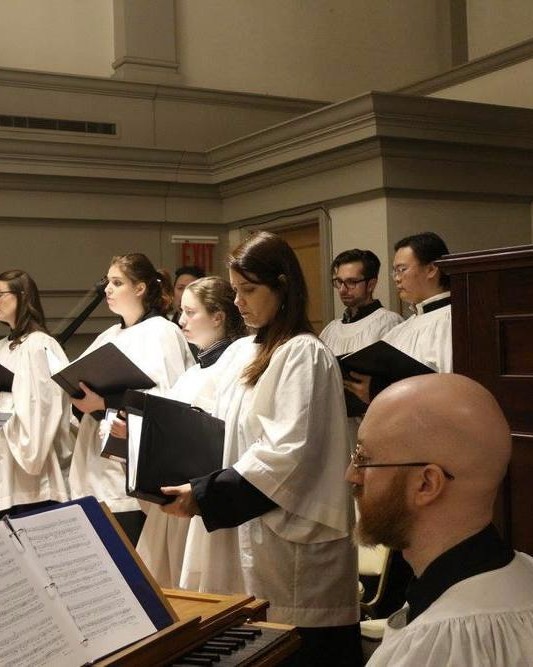 Singers in white surplices singing from scores in St. Agnes, an organist at the keyboard